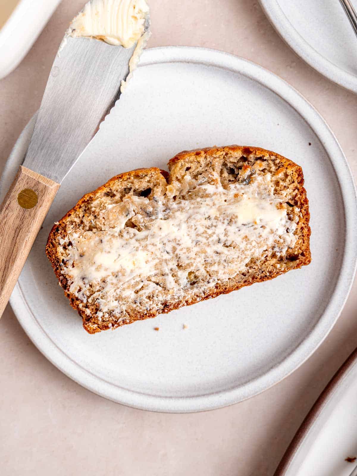 A slice of banana bread covered in butter on a plate with a butter knife.