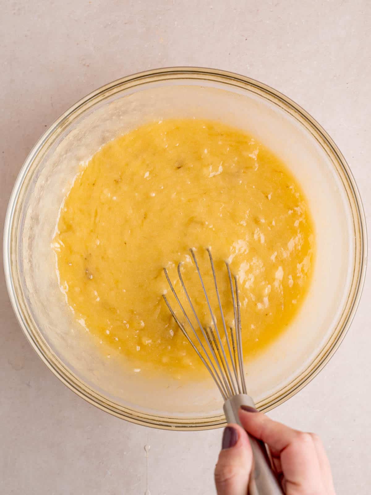 Mixing mashed bananas, melted butter, sugar, and eggs in a bowl with a whisk.