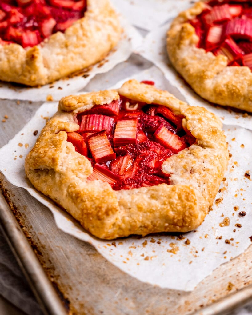 Mini strawberry rhubarb galettes baked to a golden crisp, on top of parchment paper on a metal sheet pan