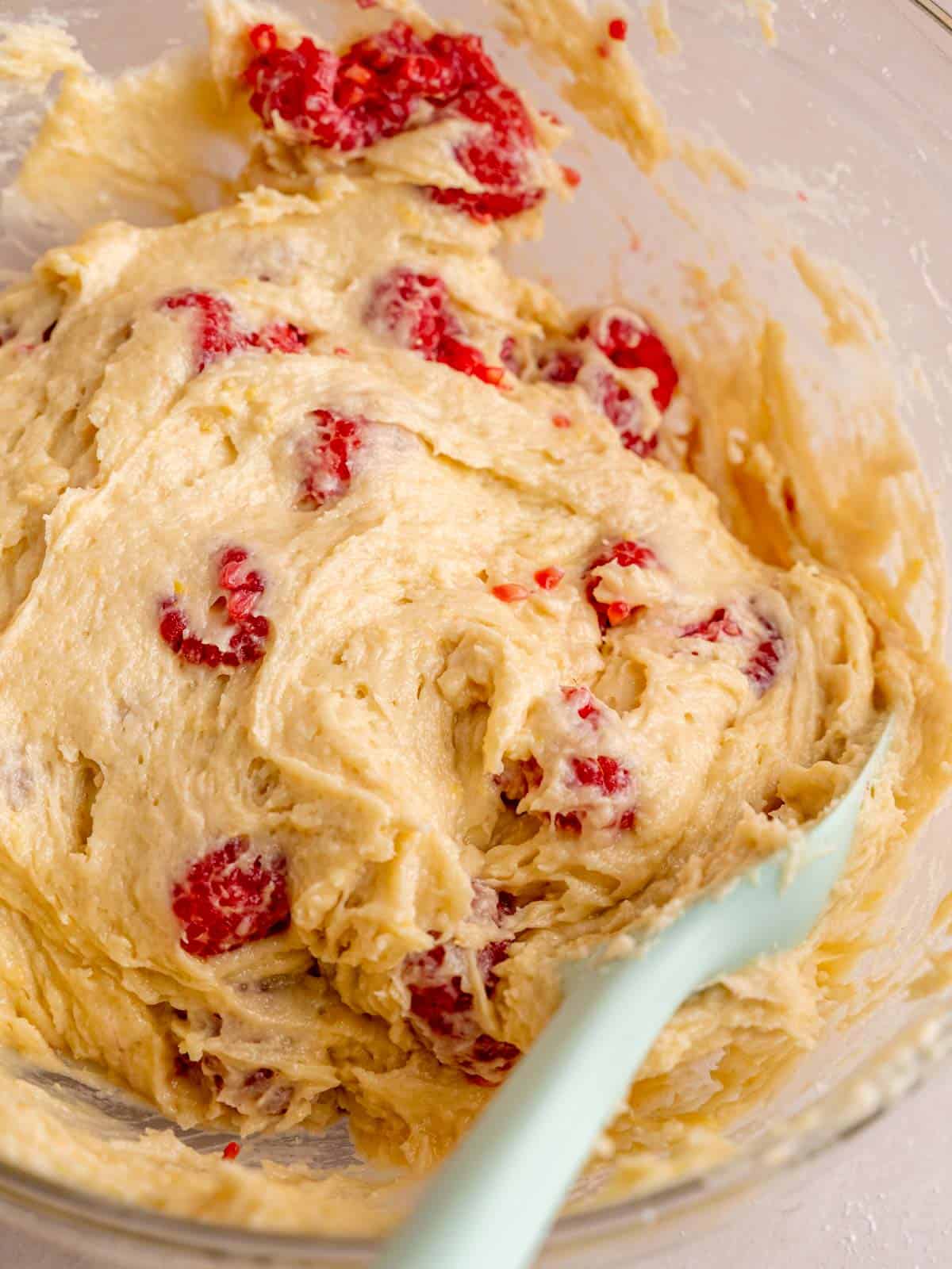 Folding fresh raspberries into lemon muffin batter using a spatula.