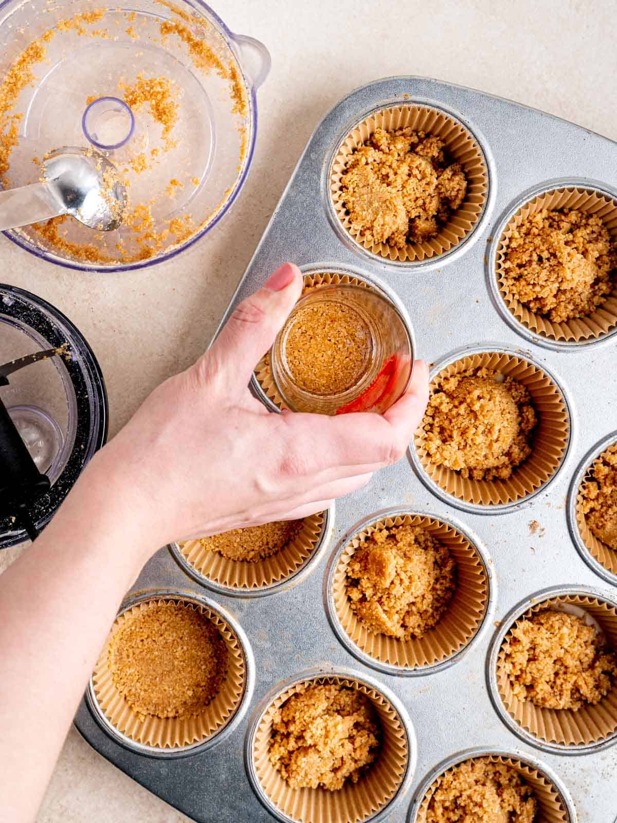 Pressing graham cracker crust into cupcake liners in a pan using the bottom of a cup.