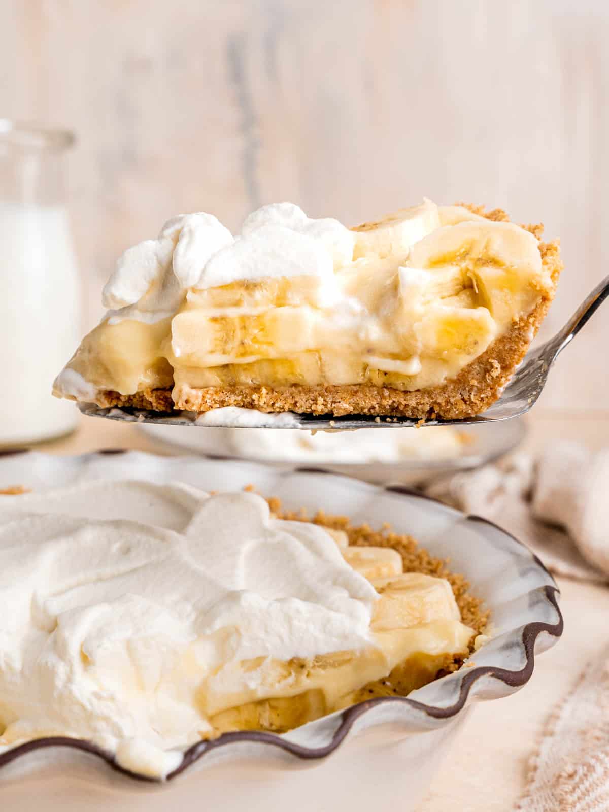 A cake server lifting a slice of banana cream pie with graham cracker crust.