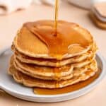 Pouring maple syrup on top of a stack of fluffy cinnamon honey pancakes.