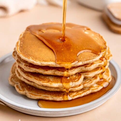Pouring maple syrup on top of a stack of fluffy cinnamon honey pancakes.