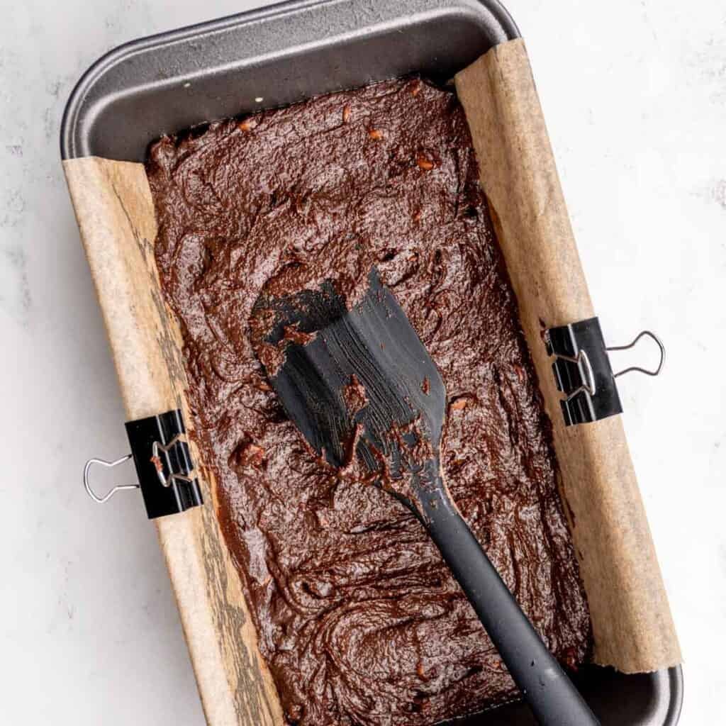 Spreading chocolate chip brownie batter into a loaf pan using a spatula.