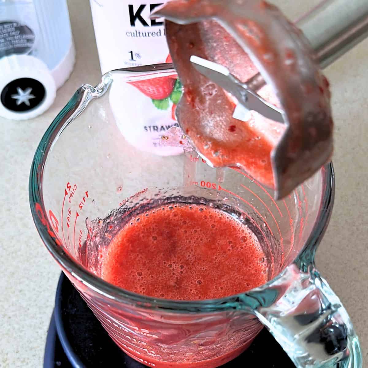 Blending warm strawberries with sugar in a measuring cup with an immersion blender.