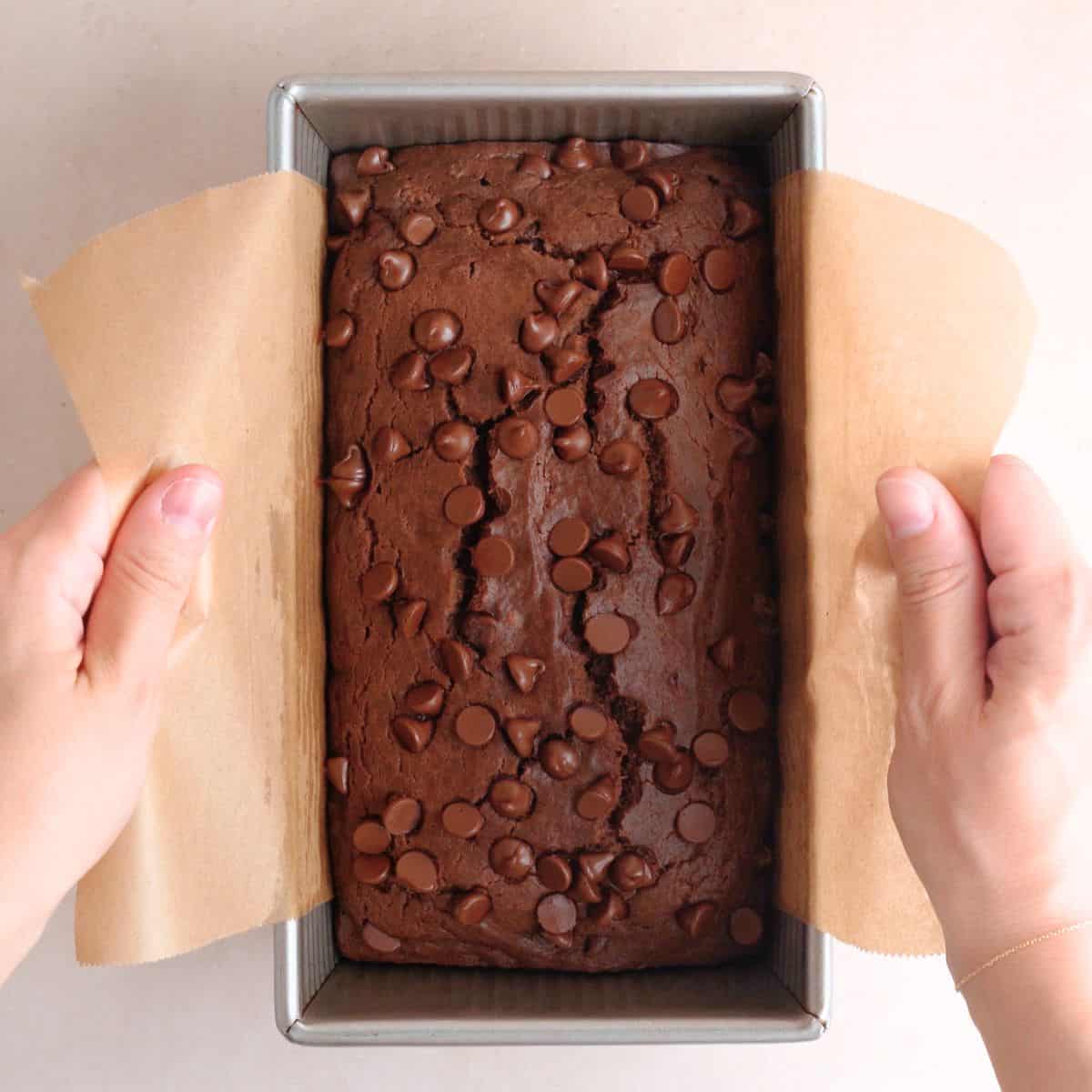 Lifting a chocolate cake out of a loaf pan with the parchment paper flaps.