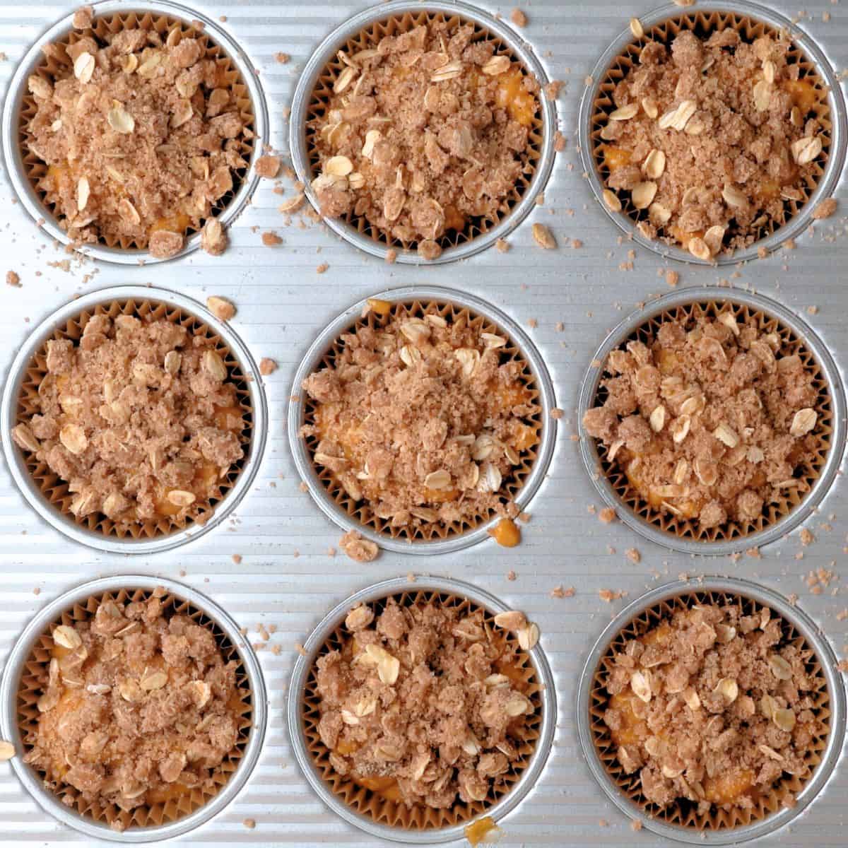 A muffin tin with oat streusel topping sprinkled over the pumpkin muffin batter.