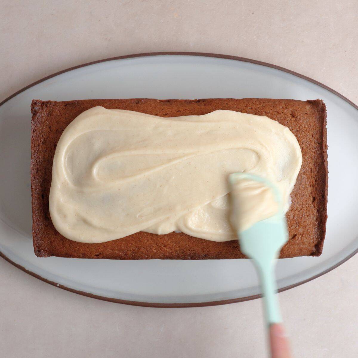 Brown butter cream cheese frosting being spread on cooled spice cake using a rubber spatula.